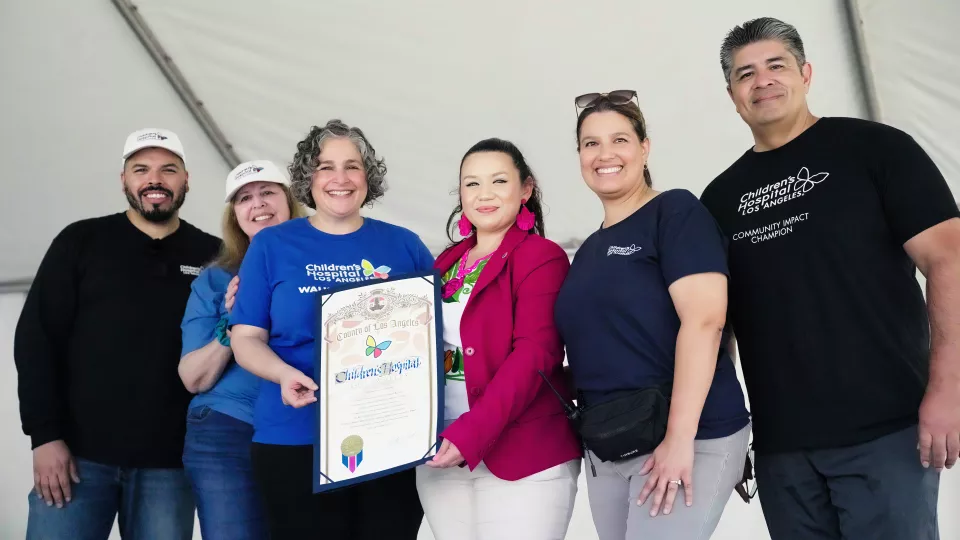 Six casually dressed adults pose with framed certificate under canopy