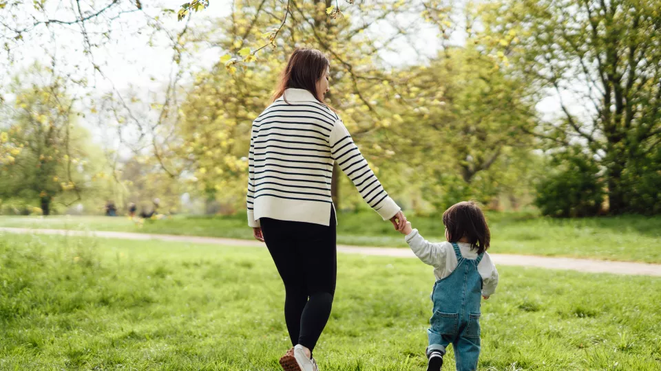 A mother holds the hand of her toddler daughter as they walk across green grass in a park