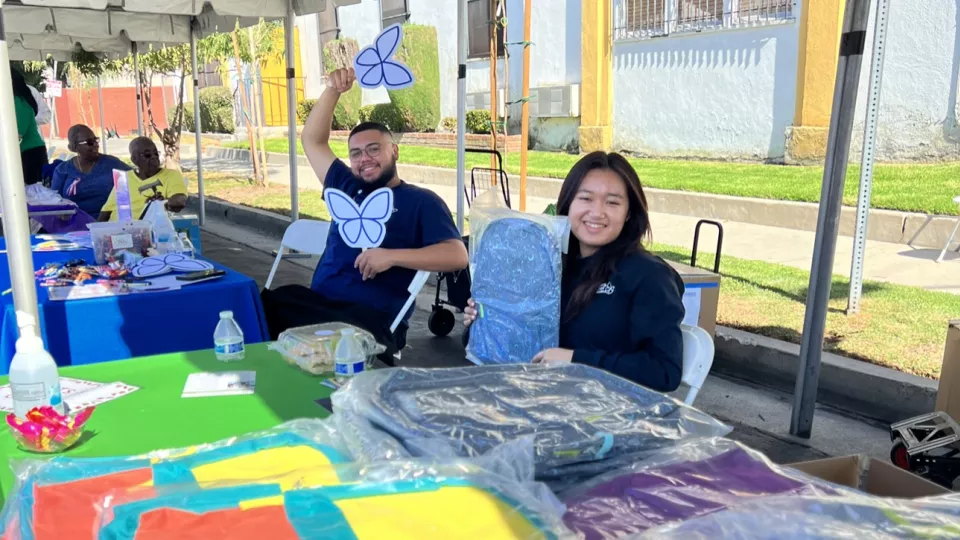 A man with medium skin tone holds up two butterfly signs, sitting next to a woman of medium skin tone holding a backpack. They sit behind a table filled with school supplies.