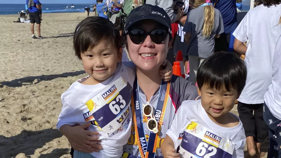 Mother with light skin tone wearing sunglasses and a cap hugs her young children on the beach