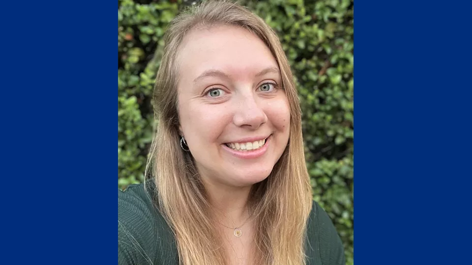 Headshot of a smiling young woman with light skin tone and long blonde hair wearing a green top against a green hedge background