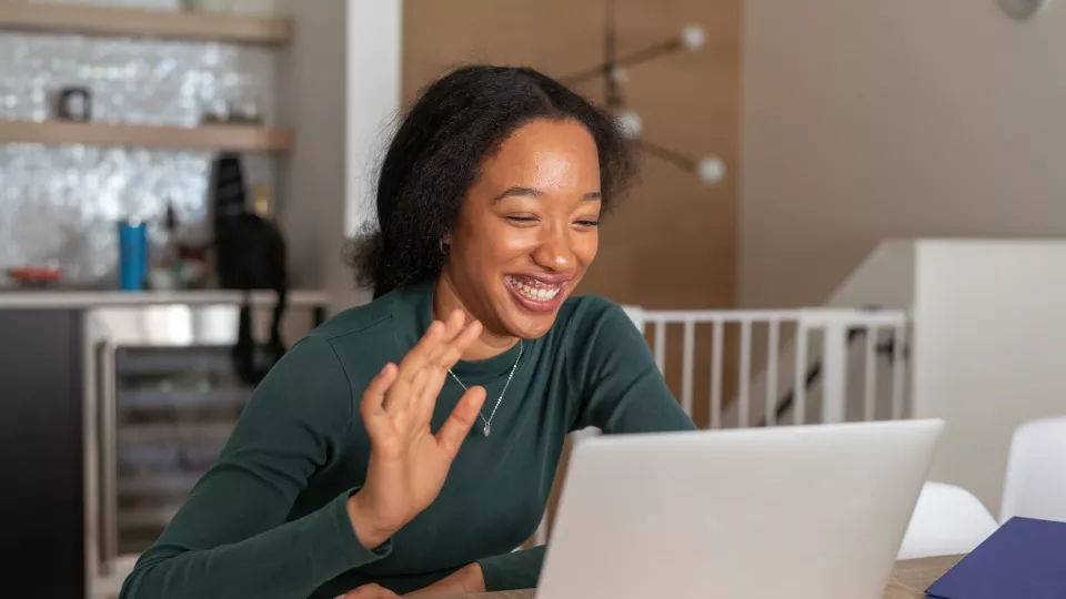 A young woman with medium skin tone and dark hair smiles and waves at her laptop screen while on a video call. She is at home