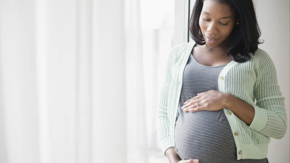 A pregnant woman with medium skin tone stands by a window and looks down while cradling her belly with both hands