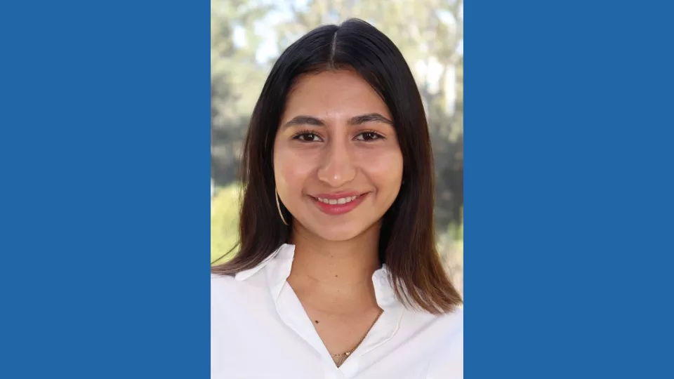 Headshot of a smiling young woman with medium skin tone and shoulder-length brown hair wearing a white blouse against a blurred outdoor background