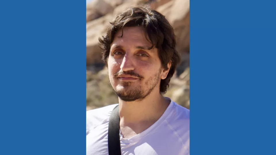 Headshot of a man with medium skin tone, brown hair and close-cropped brown beard wearing a white v-neck t-shirt against a blurred rocky, desert background