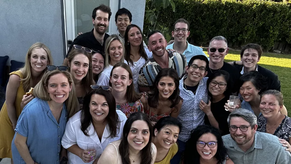 Approximately 20 casually dressed doctors smile as they pose for a group photo in a sunny backyard