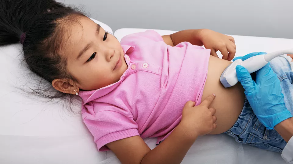 Young girl with medium skin tone and dark hair lies on her back and pulls up her pink shirt so a gloved health care provider can use ultrasound to scan her lower abdomen