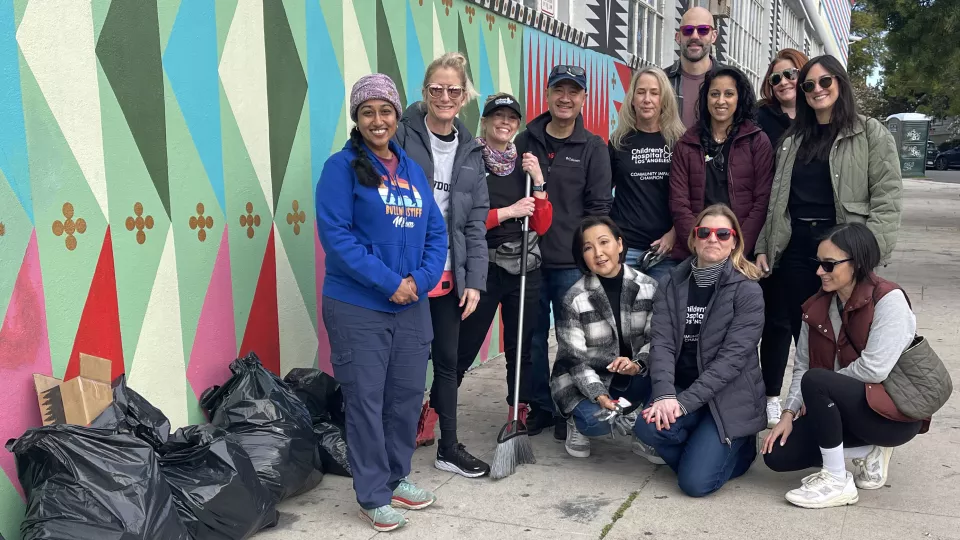 A group of men and women pose on a sidewalk in front of a colorful wall, near bags of trash.
