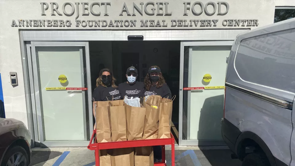 Three women in hats and surgical masks stand behind a cart filled with paper bags of food. Above them, a sign reads: Project Angel Food - Annenberg Foundation Meal Delivery Center.