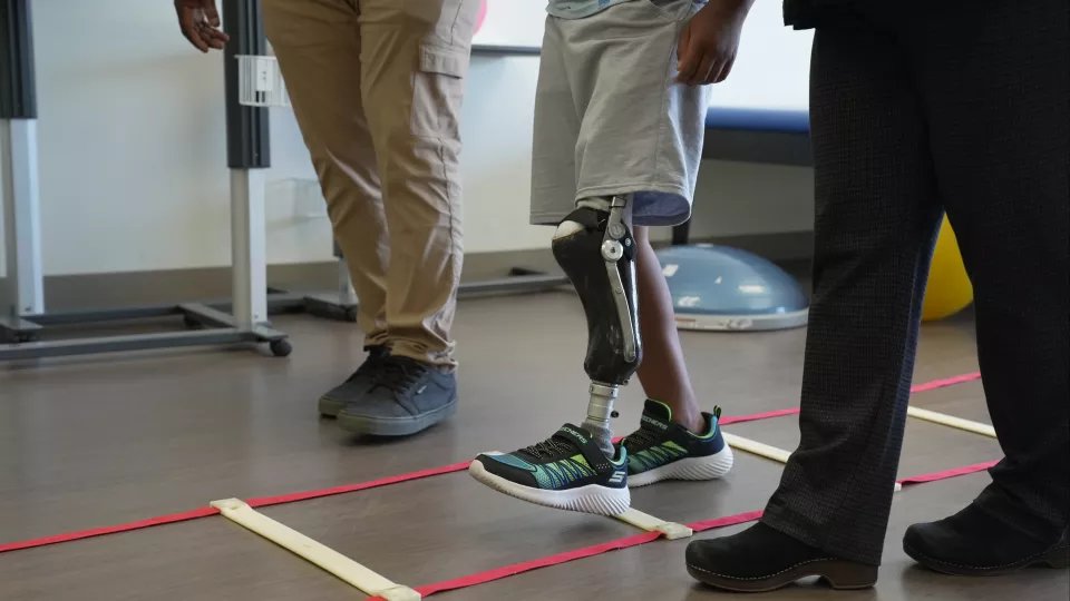 A medium-skinned boy with a prosthesis on one lower leg walks through an agility ladder on the floor. Two adults walk with him.