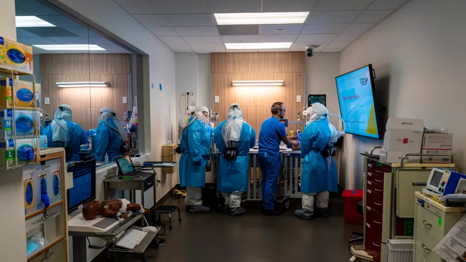 Four trainees wearing full HAZMAT suits work in a simulated hospital room while receiving guidance from an instructor