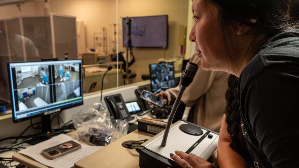 Simulation room instructor monitors trainees on quad-split computer screen and prepares to address them using a microphone