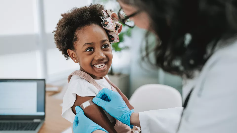 A dark skin-toned child smiles at the health care provider who is placing a bandage on her arm.