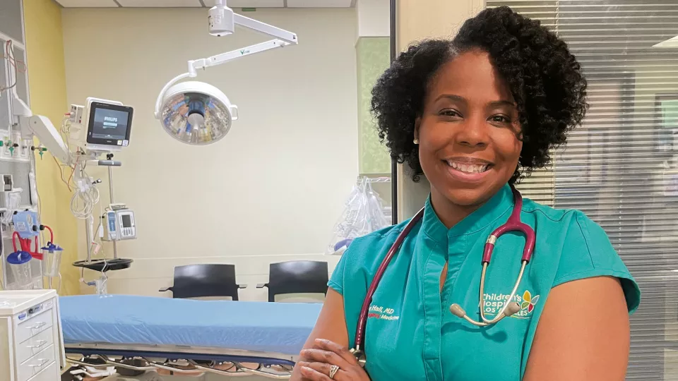 Female doctor with dark skin tone and dark hair smiles with arms crossed in hospital room