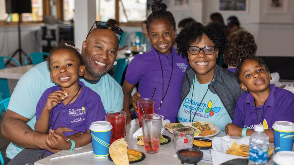 Husband and wife with dark skin tone smile as they pose with their three daughters at a restaurant table