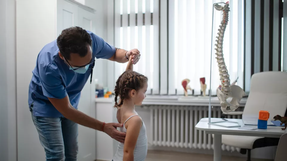 A doctor raises a girl’s left arm while checking her back in an exam room. A 3D model of the spine is in the background.