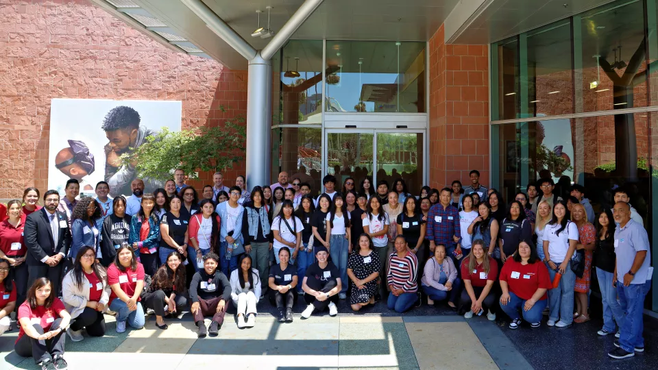 Group shot of dozens of high school-age kids and adults with a variety of skin tones, posing together on an outdoor patio outside a modern brick and glass building on a sunny day.