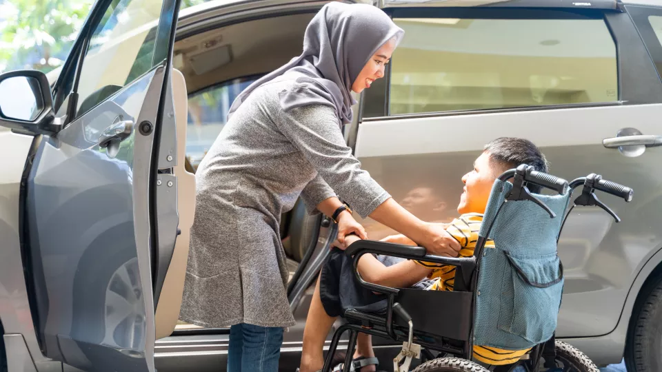 A young boy with a medium skin tone sitting in a wheelchair and smiling while an adult woman with a medium skin tone steps out of a car and smiles at him, holding his hand.