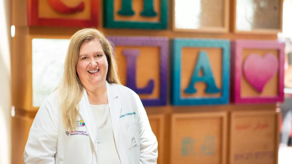 A smiling Dr. Cynthia-Herrington wears a white CHLA branded lab coat and stethoscope while standing in front of a colorful sculpture of oversized toy blocks.