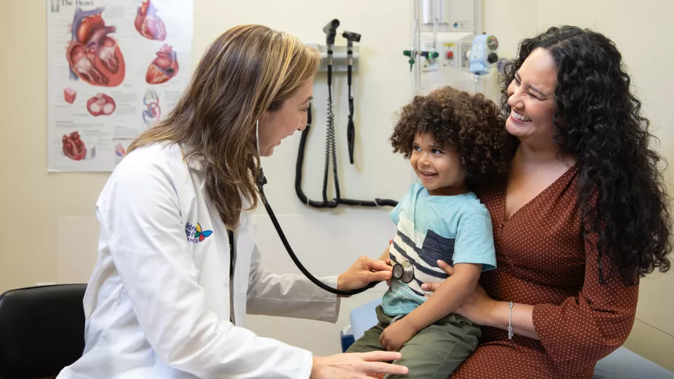 A female doctor places a stethoscope on the stomach of a young boy with curly hair. He is sitting on his mom’s lap and smiling. 