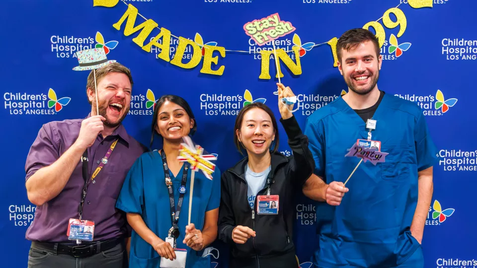 Four nurses stand and smile in front of a blue CHLA backdrop and a "Made in 1999" sign. 