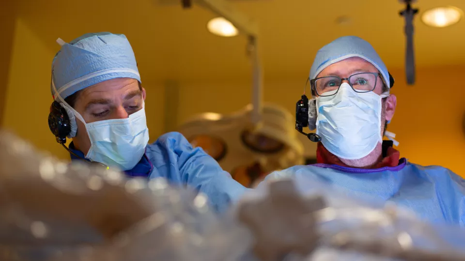 Two men in blue surgical caps, gowns and masks work side by side on a cardiac catheterization case. One is looking down at an unseen patient.