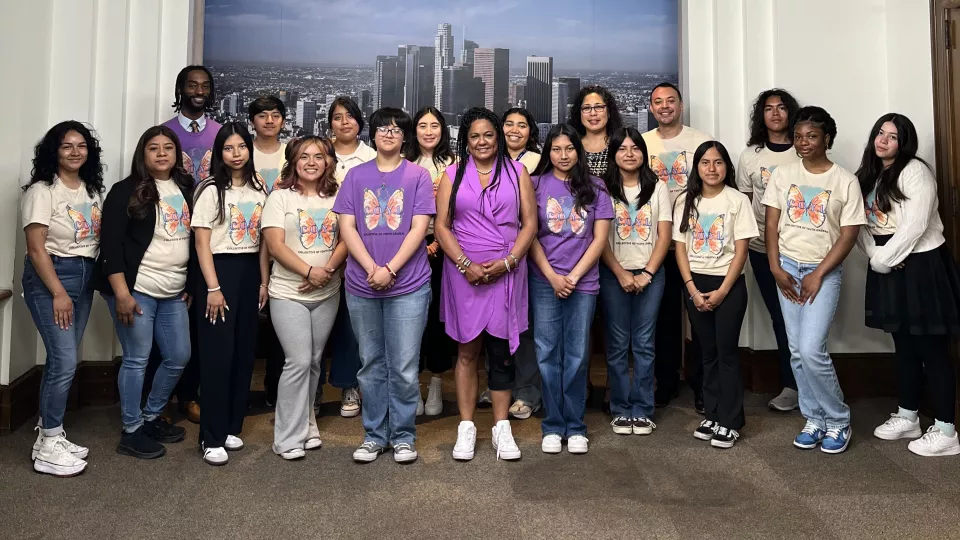 Group shot of high school-age kids with medium, medium-dark and dark skin tones posing with an adult man and an adult woman with a medium skin tone, and an adult man with a dark skin tone, all standing indoors in front of a large photo of the Los Angeles skyline.