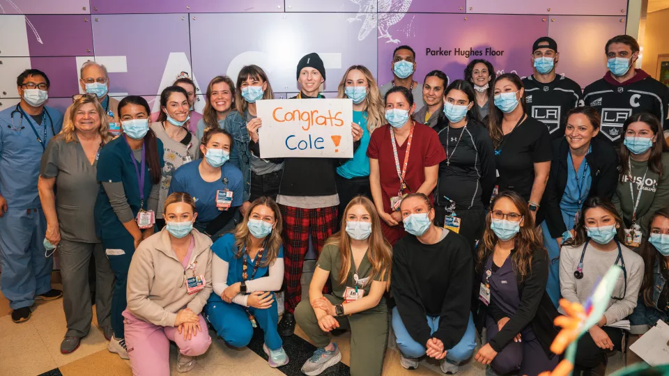 Group photo of teenage boy with light skin tone who is smiling as he holds sign reading "Congrats Cole" and is surrounded by members of his masked cancer care team