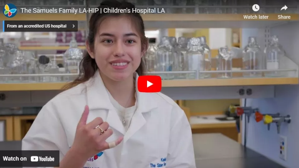 Young woman with light skin tone wearing white lab coat smiles as they sit in a laboratory setting in a screengrab of a YouTube video player