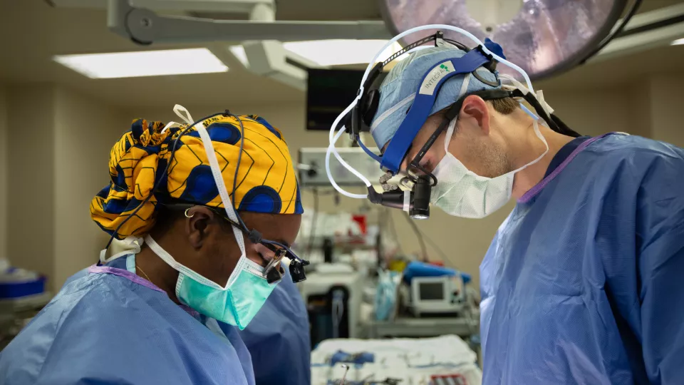 A dark-skinned female surgeon with a gold-and-blue surgical cap and a light-skinned male surgeon look down at an unseen operating table.