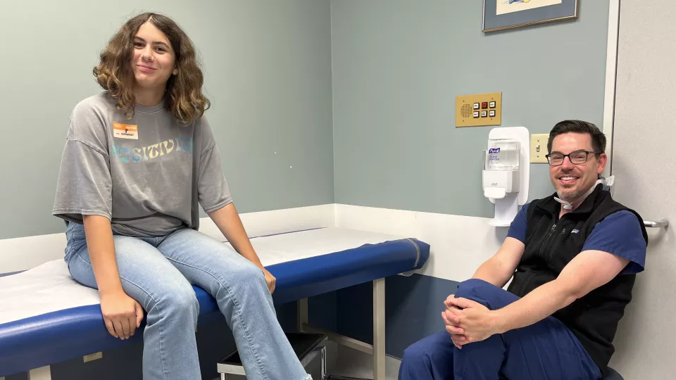 Teenage girl in blue jeans seated alongside medical driver sitting on stool