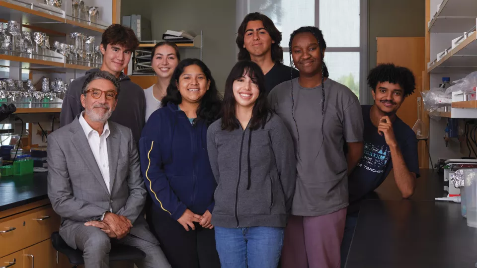 Chuck Lorre, a man with light skin and grey hair, wearing a white shirt and grey suit sits with seven casually dressed high school students in a laboratory setting