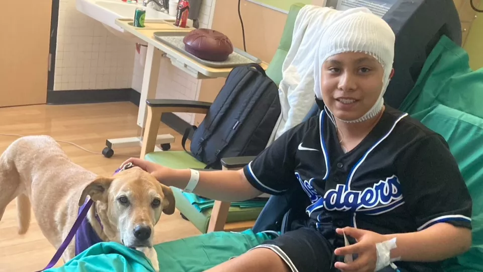 Young boy in hospital room bed with bandaged head and Dodgers jersey and dog at bedside