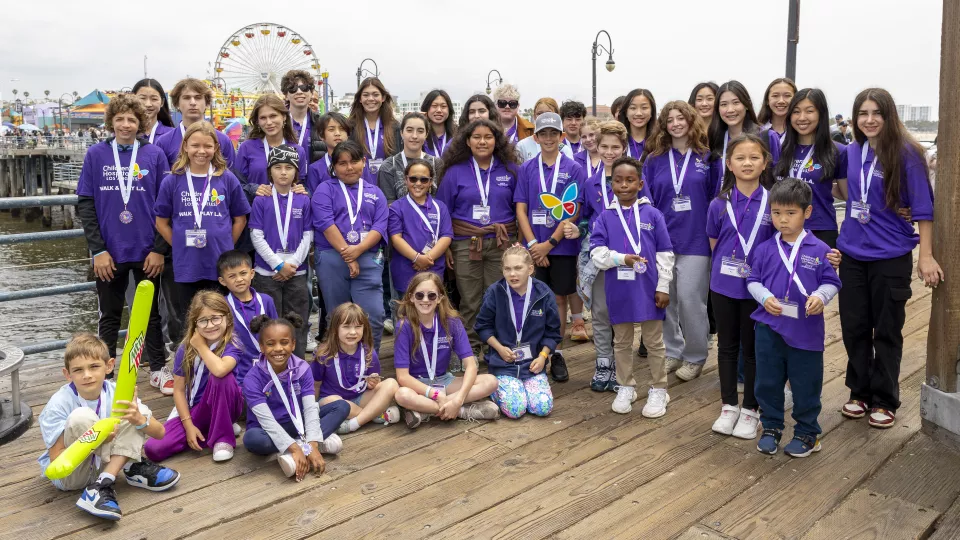 Around 30 children and teenagers wearing matching purple Children's Hospital Los Angeles polo shirts pose for a team photo on the Santa Monica Pier