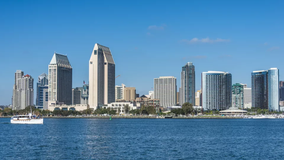 An array of tall buildings on the shore of a blue bay, against a clear blue sky. A white boat is in the water.