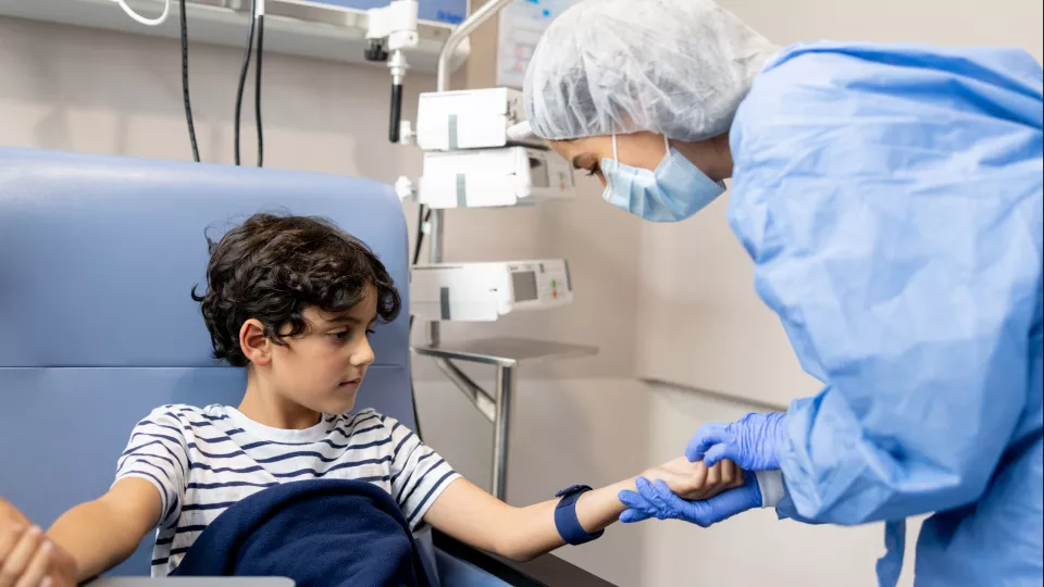 A female nurse in a blue surgical gown and gloves holds the arm of a young boy with dark curly hair, looking for a vein.