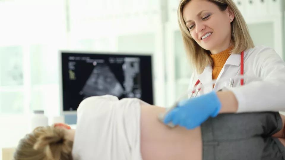 A smiling female doctor places an ultrasound probe on a child’s back. An ultrasound image is on a screen in the background.