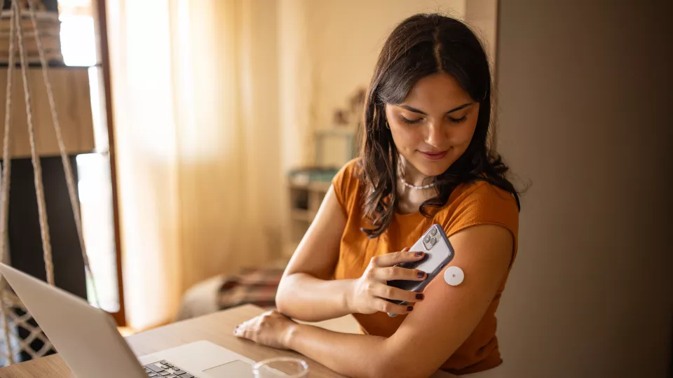 A medium-skinned young woman with long dark hair holds her smartphone over the continuous glucose monitor on her arm.