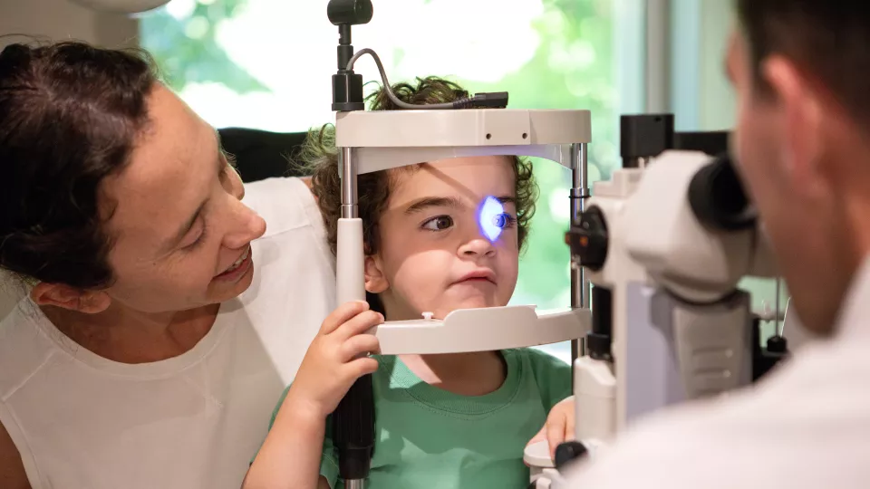 A light shines in a young child’s eyes as an unseen ophthalmologist checks her vision. The child is sitting on her mother’s lap.