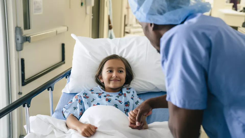 A medium-skinned girl on a gurney smiles at a dark-skinned doctor in blue scrubs and surgical cap, who is holding her hand.