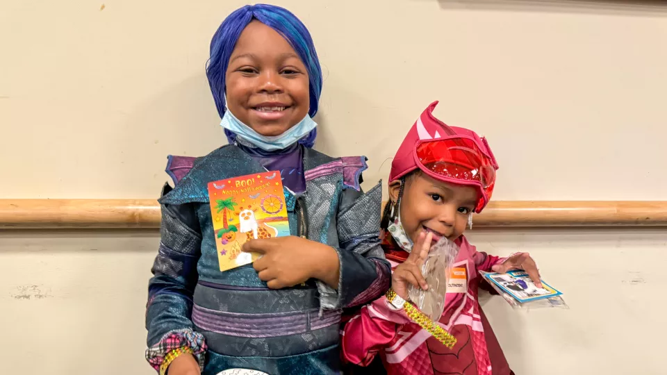 Two young sisters dressed in superhero Halloween costumes hold up their CHLA Halloween cards. 