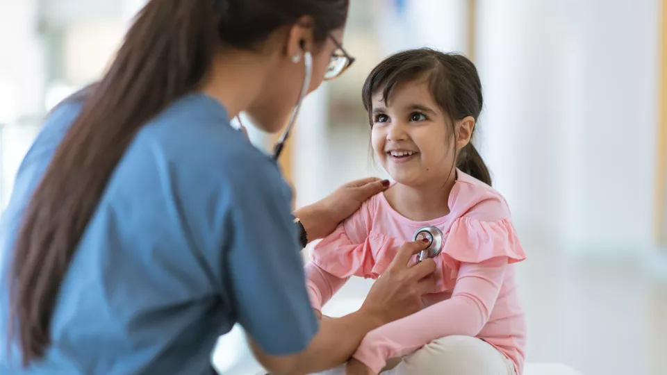 Woman in blue scrubs uses a stethoscope to listen to a child’s heartbeat.