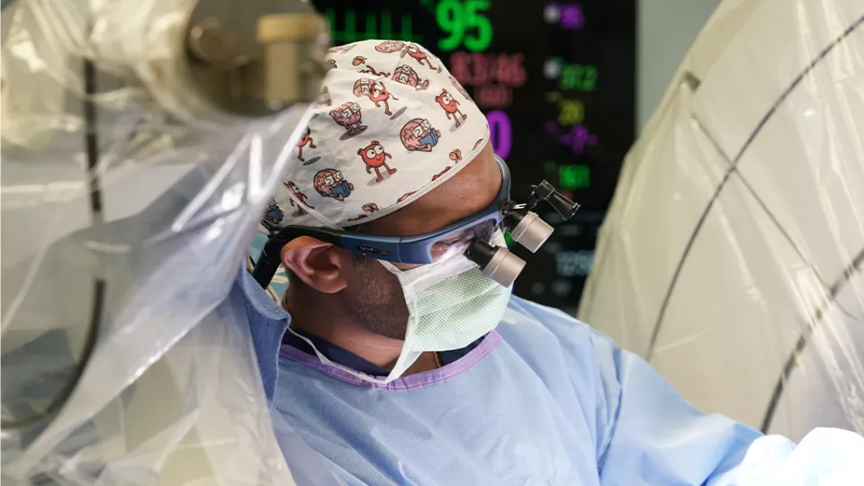 A man wearing blue scrubs, surgical loupes, mask, and a white surgical cap with colorful characters on it focuses on a target amidst machinery covered in plastic