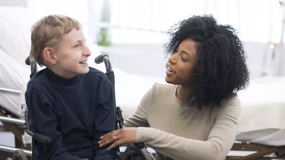 A light-haired boy in a wheelchair smiles at a dark-haired woman who is crouched down beside him in front of a white hospital bed.