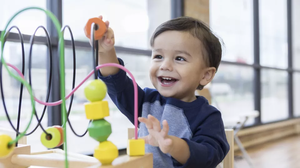 A small boy with medium-light skin tone smiles while moving blocks along a wire toy. He is sitting near bright windows.