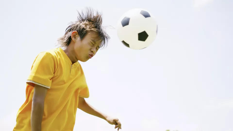 Boy in a bright yellow short-sleeved shirt closes his eyes and winces as he hits a soccer ball with his head.