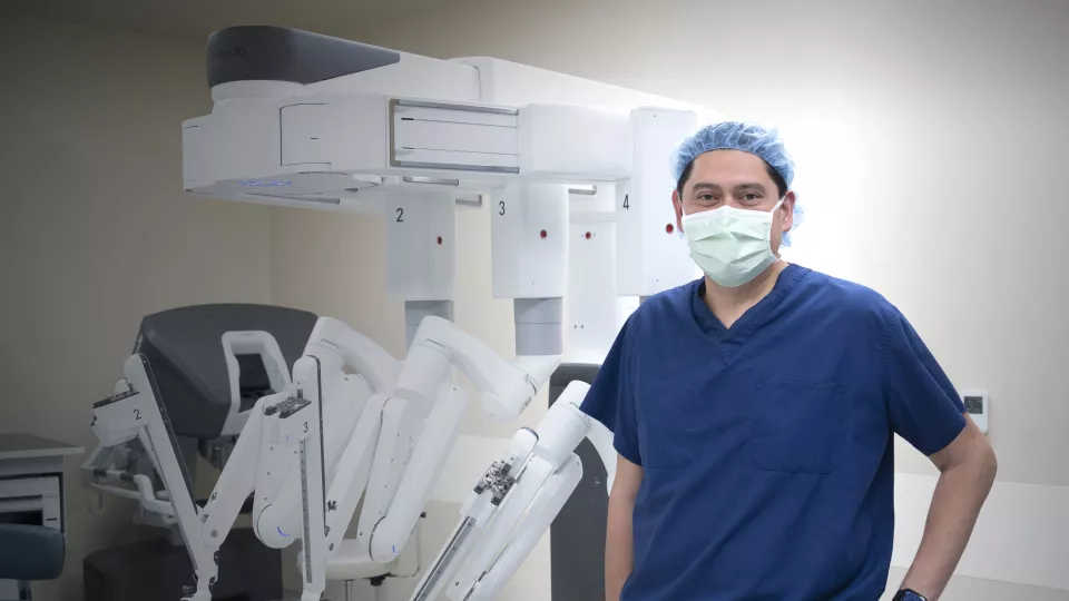 A man with medium skin tone in dark blue scrubs and a light blue surgical cap and mask stands in front of a large white machine with four arms