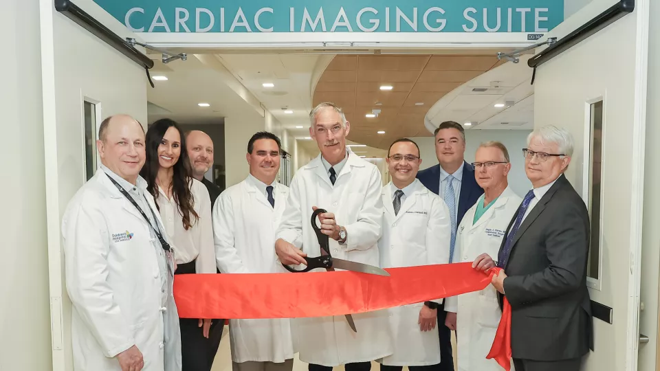 A physician in a white lab flanked by colleagues on either side uses an oversized pair of scissors to cut a red ribbon in a hospital corridor