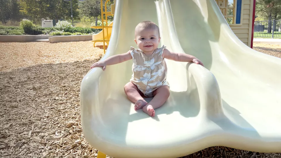 Toddler with medium skin tone smiles as they sit at the bottom of a playground slide on a bright, sunny day