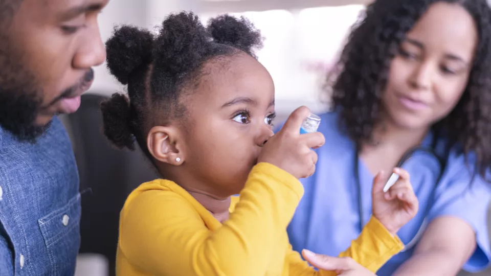 A young girl sits between her parents while using an inhaler for asthma. 